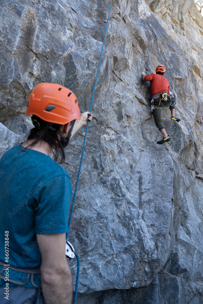 Young man climbing on rock while his colleague holds the safety harness Young man climbing on rock while his colleague holds the safety harness