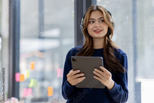 Young adult happy smiling Hispanic Asian student wearing headphones standing indoor using digital tablet in university campus or at virtual office. College female student learning remotely.