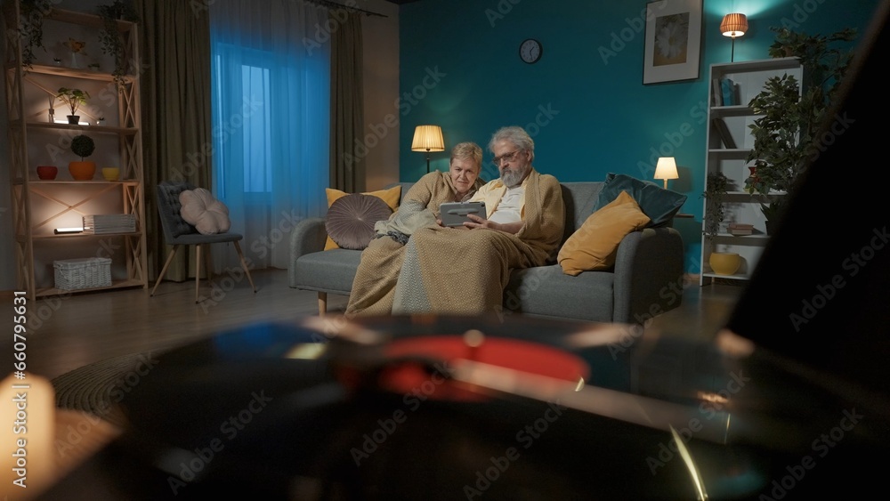 Close up shot shows a vinyl record player with a vinyl record. In the background an elderly couple is sitting on a couch with a tablet looking at something, web sites or social media Close up shot shows a vinyl record player with a vinyl record. In the background an elderly couple is sitting on a couch with a tablet looking at something, web sites or social media
