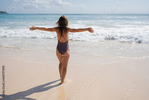 Cheerful female in swimsuit running in foamy ocean near sandy beach in sunny day. 