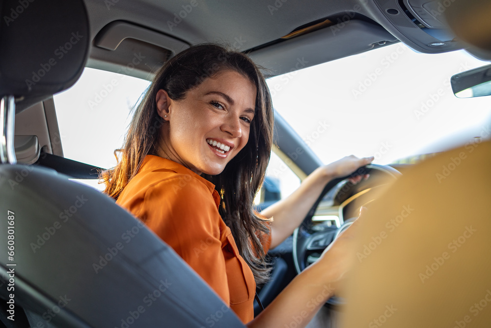 Young woman sitting on a driver's seat in the car and looking at camera over the shoulder. Portrait of pleasant female with positive expression, being satisfied with unforgettable journey by car. Young woman sitting on a driver's seat in the car and looking at camera over the shoulder. Portrait of pleasant female with positive expression, being satisfied with unforgettable journey by car.