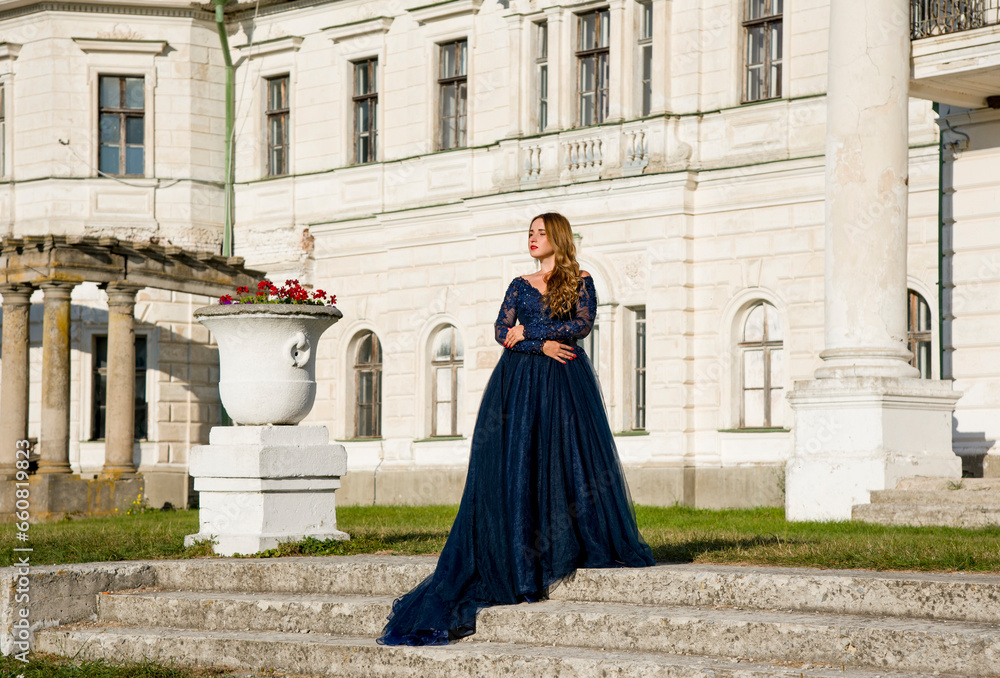 Beautiful brunette woman with curly hair and red lips in long blue dress walking near big white palace with columns. Princess in palace conception