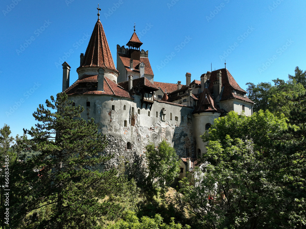 Bran Castle in Transylvania, Romania, unfolds with its distinctive architecture. Nestled amidst the natural canopy against the backdrop of a clear blue sky, the fortress embodies a timeless allure.