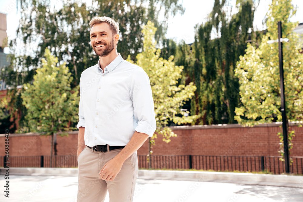 Handsome smiling stylish hipster lambersexual model. Modern man dressed in white shirt and trousers. Fashion male posing in the street background