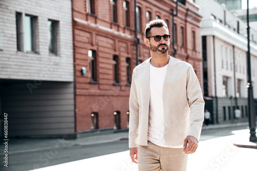 Portrait of handsome confident stylish hipster lambersexual model. Sexy modern man dressed in elegant suit. Fashion male posing in the street background in Europe city at sunset. In sunglasses