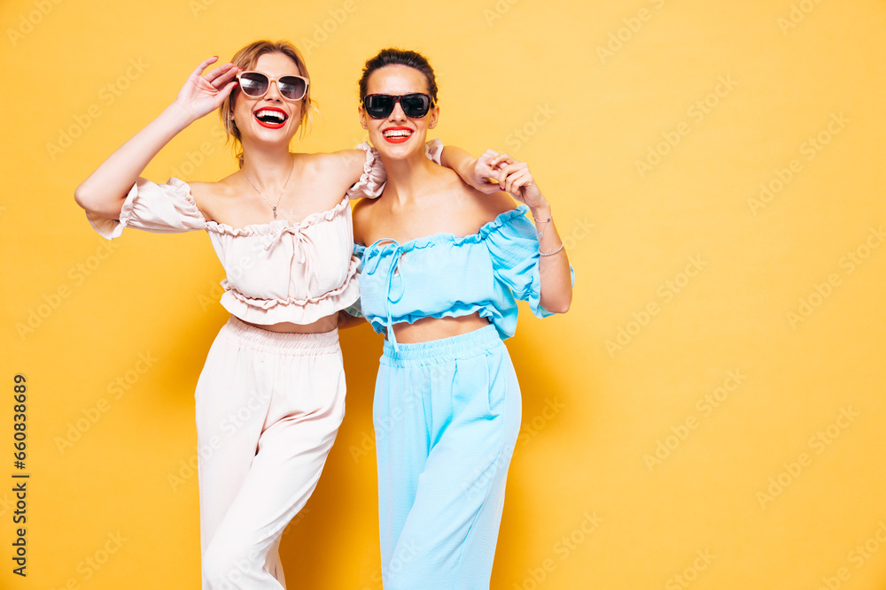 Two young beautiful smiling brunette hipster female in trendy summer clothes. Sexy carefree women posing near yellow wall in studio. Positive models having fun. Cheerful and happy. in sunglasses