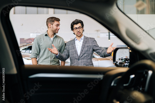 View from interior of luxury vehicle to professional car dealer in business suit showing new car to cheerful male client explaining characteristics. Bearded salesman helping with choice of auto to guy © dikushin