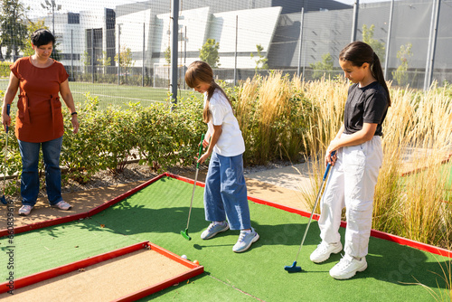 Cute school girl playing mini golf with family. Happy toddler child having fun with outdoor activity. Summer sport for children and adults, outdoors. Family vacations or resort. photo