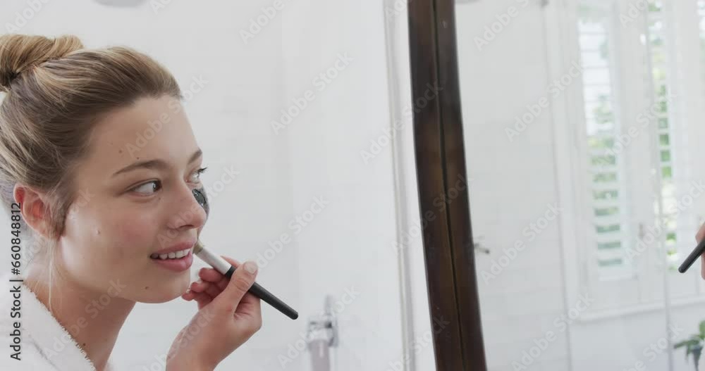 Happy biracial woman applying beauty face mask looking at mirror in bathroom, slow motion