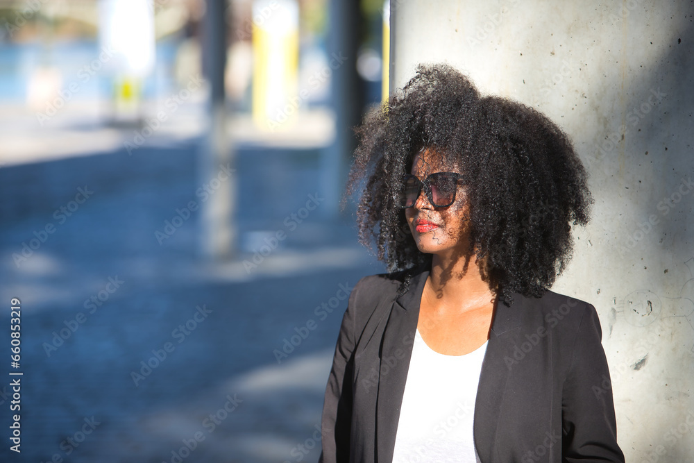 Young, beautiful, black woman with afro hair, jacket and sunglasses leaning on a concrete column receiving the sun's rays. Concept empowerment, success, current, modern.