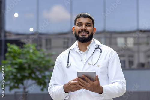 Portrait of a young Muslim male doctor standing in a white coat near a clinic on the street and holding a tablet in his hands, smiling and looking at the camera © Tetiana