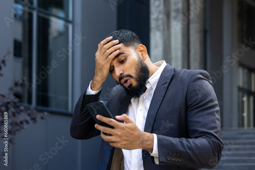 Upset Muslim male businessman standing near office and holding head, shocked reading news, message received