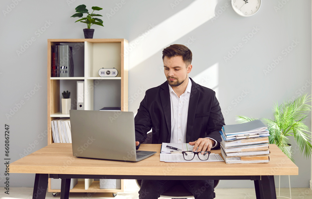 Fototapeta premium Focused businessman working at home with laptop and papers on desk. Portrait of handsome young man in formal wear sitting at desk in office and looking at laptop screen