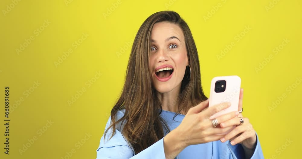 Attractive brunette curly woman look surprised wow hold using mobile cell phone on yellow background. Girl look on smartphone and doing winner gesture. Girl just found out big win news, point on phone Attractive brunette curly woman look surprised wow hold using mobile cell phone on yellow background. Girl look on smartphone and doing winner gesture. Girl just found out big win news, point on phone