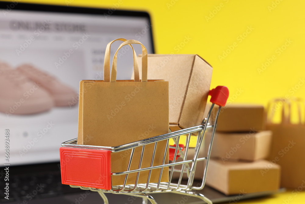Online store. Mini shopping cart, purchases and laptop against yellow background, selective focus