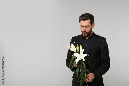 Sad man with white lilies mourning on light grey background, space for text. Funeral ceremony