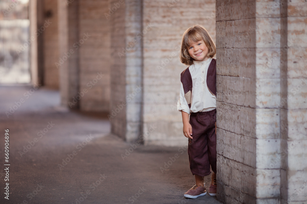 Little boy with long hair in brown clothes on city street in summer 