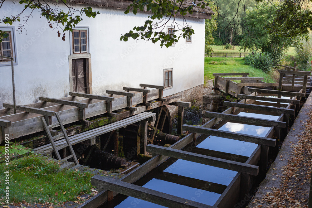 View of a water mill system. The mill's wheels and water pipes are visible, and the water reflects the sky.