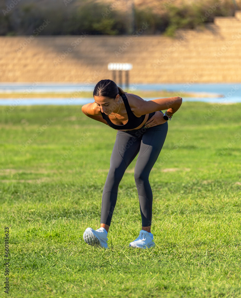 Beautiful young runner girl, tanned with long dark hair, leaning towards her leg with her hands on her waist to warm up and stretch her muscles before training. Beautiful young runner girl, tanned with long dark hair, leaning towards her leg with her hands on her waist to warm up and stretch her muscles before training.