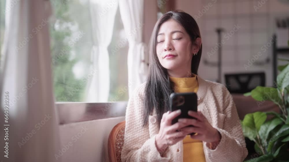 Portrait of attractive asian woman holding smartphone using app on Internet sitting in chair at home looking outside window and smiling dreamy Portrait of attractive asian woman holding smartphone using app on Internet sitting in chair at home looking outside window and smiling dreamy