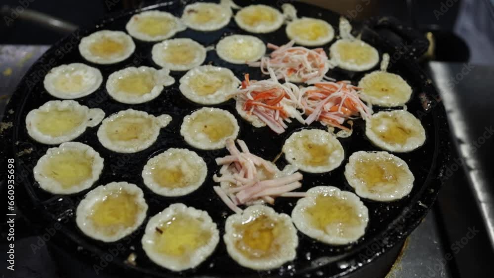 Food vendor putting ham on crispy fried mini pancakes using tongs in Thai-styled Takoyaki pan at market. Cooking crispy fried seafood mini pancakes. Street food Thailand. Fried seafood omelet pancake. Food vendor putting ham on crispy fried mini pancakes using tongs in Thai-styled Takoyaki pan at market. Cooking crispy fried seafood mini pancakes. Street food Thailand. Fried seafood omelet pancake.