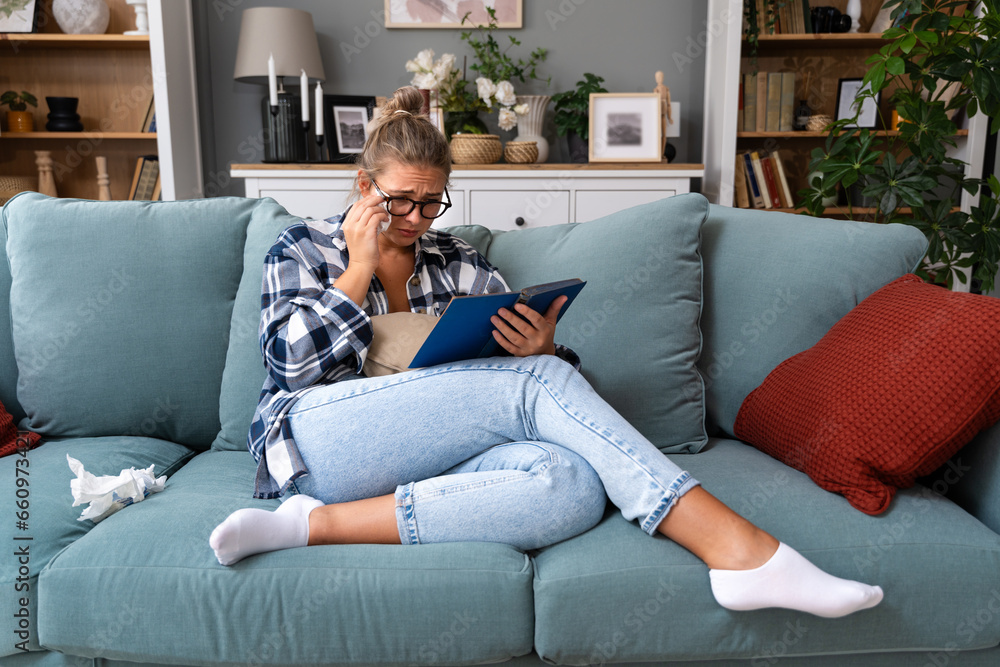 Sad woman crying while she reading a good paper book sitting on a sofa at home. Upset girl read love story cry of sadness - obrazy, fototapety, plakaty 