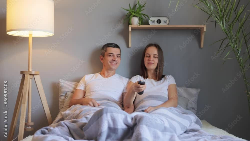 Happy relaxed young family couple lying in bed holding remote control watching tv together lounging talking enjoying television in cozy bedroom at home. Happy relaxed young family couple lying in bed holding remote control watching tv together lounging talking enjoying television in cozy bedroom at home.