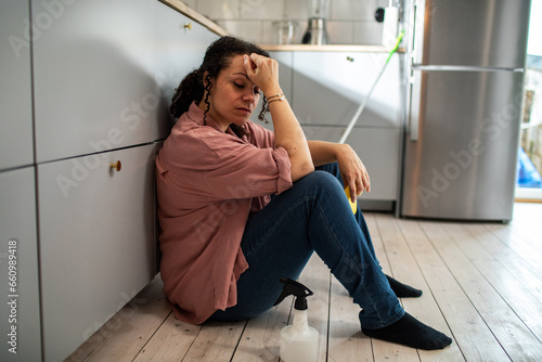 Tired young mixed woman sitting on the floor after cleaning the kitchen photo