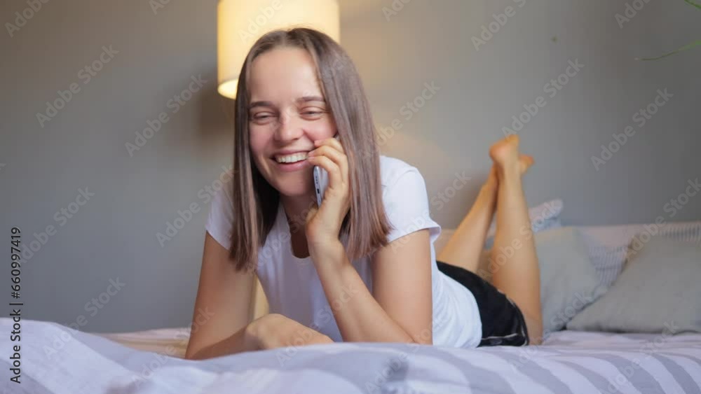Extremely happy cheerful woman lying on bed wearing pajama talking on smartphone enjoying funny conversation laughing out loud spending weekend at home with phone communication. Extremely happy cheerful woman lying on bed wearing pajama talking on smartphone enjoying funny conversation laughing out loud spending weekend at home with phone communication.