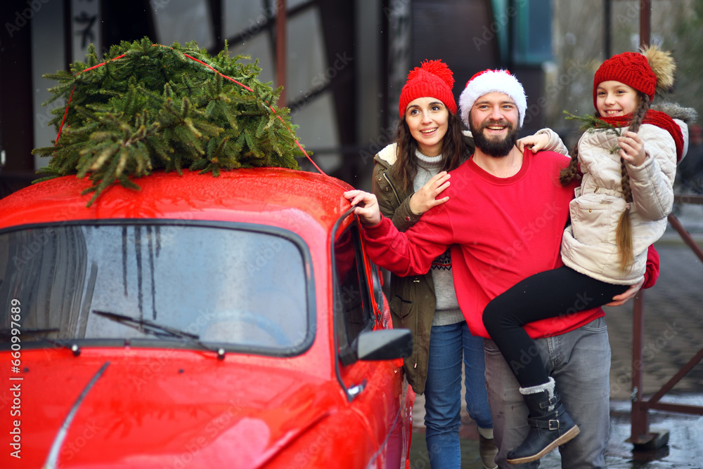 mom, dad and a daughter in a knitted red hat with pigtails hug and smile against the background of a red car with a Christmas tree on the roof mom, dad and a daughter in a knitted red hat with pigtails hug and smile against the background of a red car with a Christmas tree on the roof