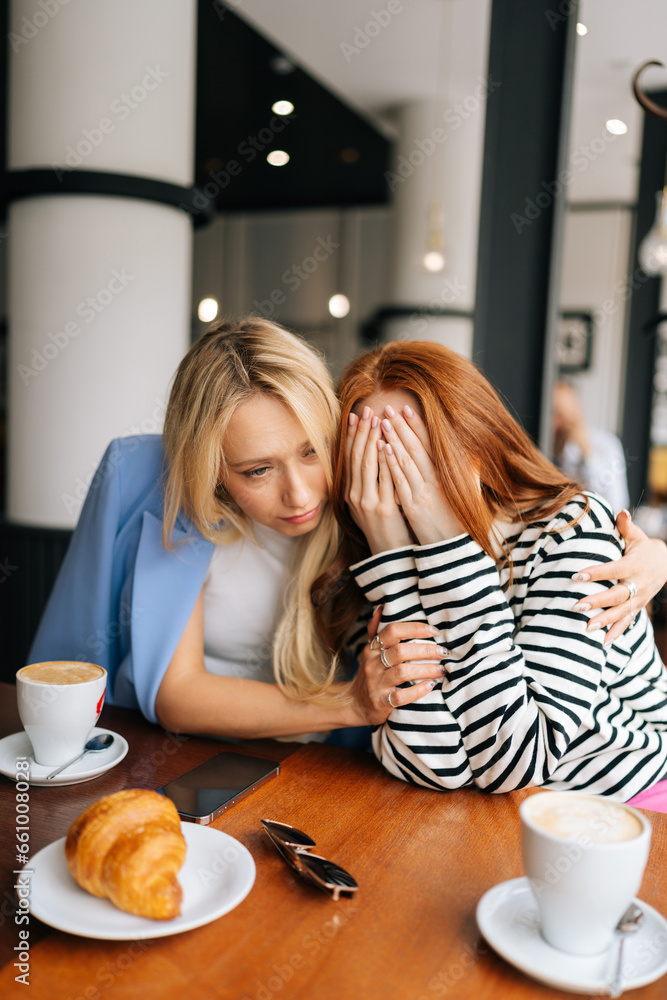 Vertical portrait of two best girlfriends sitting at indoors cafe and talking. Blonde female stroking hand, hugging, trying calm unhappy redhead friend, who cover face with hands and tearing. Vertical portrait of two best girlfriends sitting at indoors cafe and talking. Blonde female stroking hand, hugging, trying calm unhappy redhead friend, who cover face with hands and tearing.