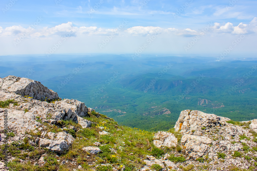 cliff of a mountain with stones, grass and flowers in the foreground, distant hills covered with forest and sky with clouds in the background in the Crimean mountains in Europe cliff of a mountain with stones, grass and flowers in the foreground, distant hills covered with forest and sky with clouds in the background in the Crimean mountains in Europe