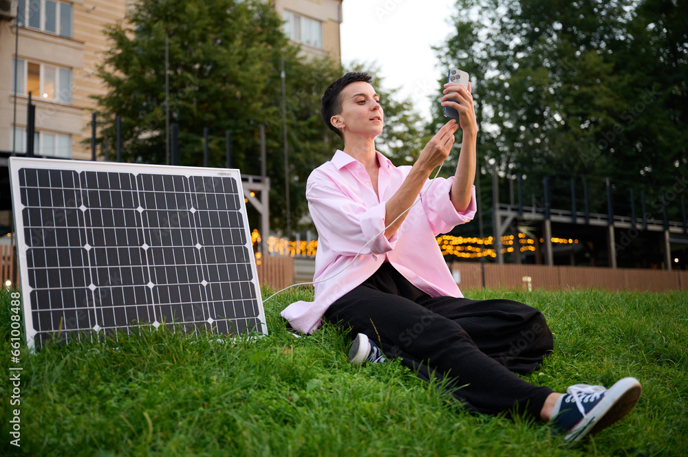 Fototapeta premium Young woman charging her phone by solar panel. Pretty female in casual clothes sitting on grass and pluging charger in her phone. Renewable energy concept.