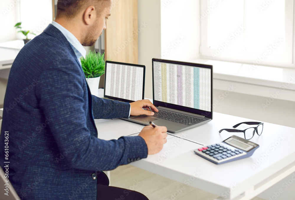 Busy concentrated male financier working in office with Excel spreadsheets and calculator. Man sits in front of two laptops, fills out paper documents and enters data into electronic files.