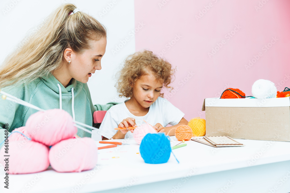 Mom teaches her daughter to knit. Mom and a little blonde girl are sitting at the table and crocheting. Mom teaches her daughter to knit. Mom and a little blonde girl are sitting at the table and crocheting.