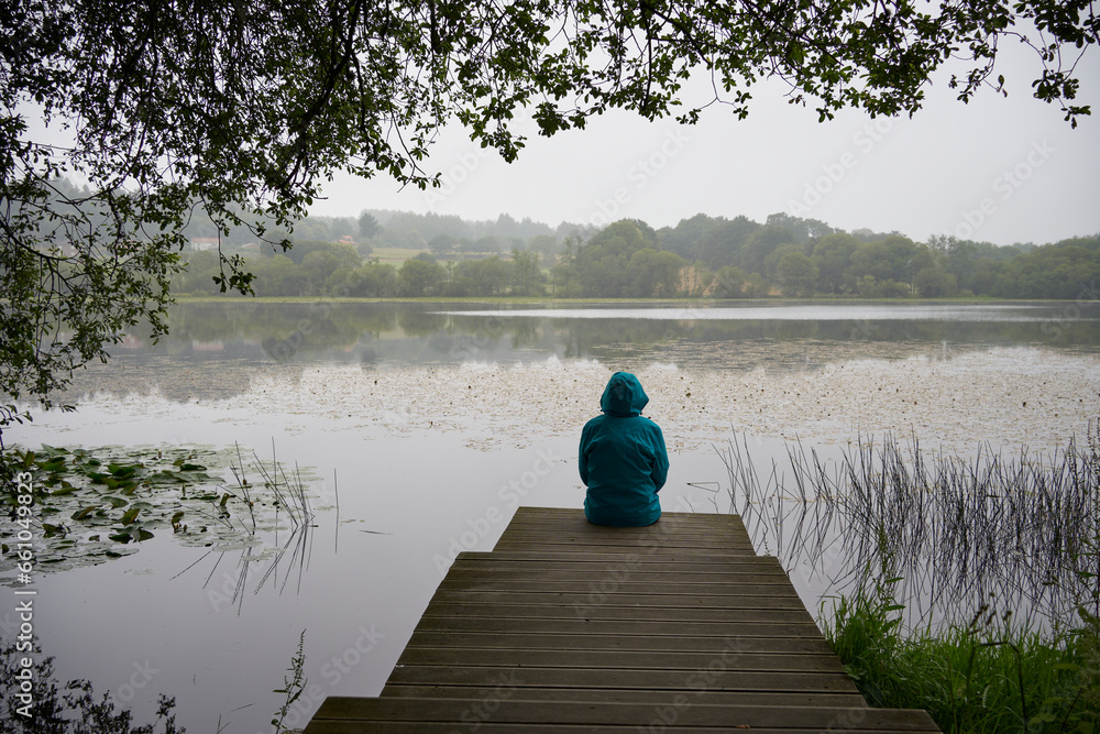 Rear view of silhouette of woman with blue raincoat sitting on the wooden pier in front of the lake on a rainy day. Concept of rural life, nature and landscape.