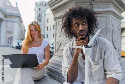 An engineering student is thoughtful as he holds a small wind turbine and his friend goes over her notes