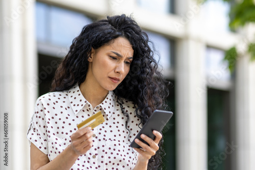 Upset and disappointed woman received money transfer error, Hispanic woman with bank credit card and phone cheated walking down street outside office building.