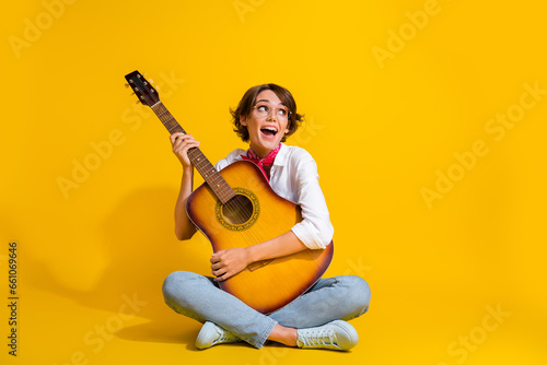 Photo of cheerful lovely girl wear trendy clothes sitting floor playing guitar looking empty space ad isolated on yellow color background