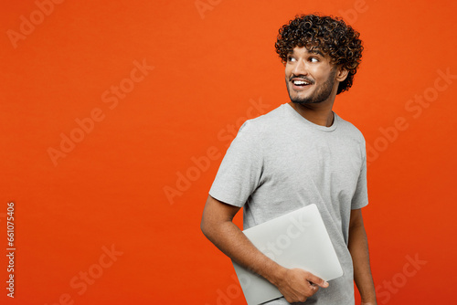 Side view young smiling fun happy Indian man wears t-shirt casual clothes hold closed laptop pc computer browsing internet isolated on orange red color background studio portrait. Lifestyle concept.
