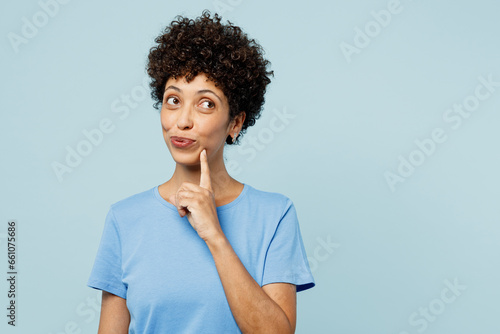 Young fun woman of African American ethnicity wear t-shirt casual clothes put hand prop up on chin, lost in thought and conjectures isolated on plain pastel light blue cyan background studio portrait.