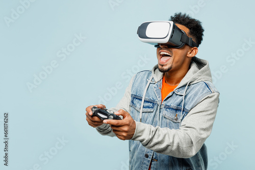 Young man of African American ethnicity in denim jacket orange t-shirt hold in hand play pc game with joystick console watching in vr headset pc gadget isolated on plain pastel light blue background