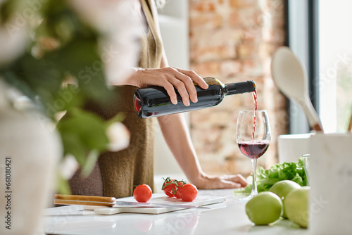 cropped woman pouring red wine into glass near fresh apples and cherry tomatoes on countertop photo