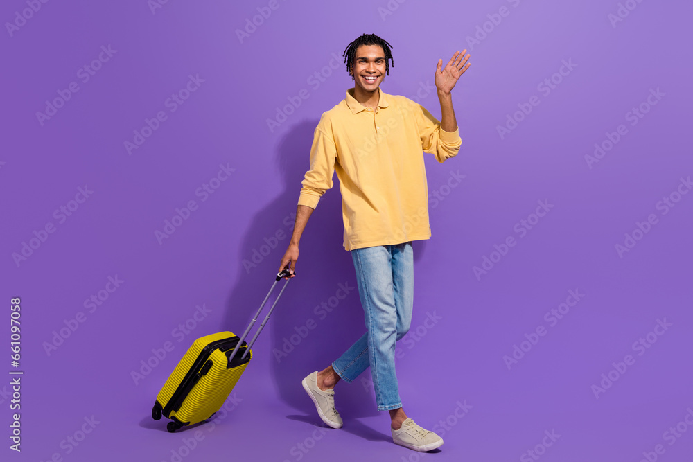 Full size body photo of funky guy greetings waving palm positive hold baggage airport traveling tourist isolated on violet color background Full size body photo of funky guy greetings waving palm positive hold baggage airport traveling tourist isolated on violet color background