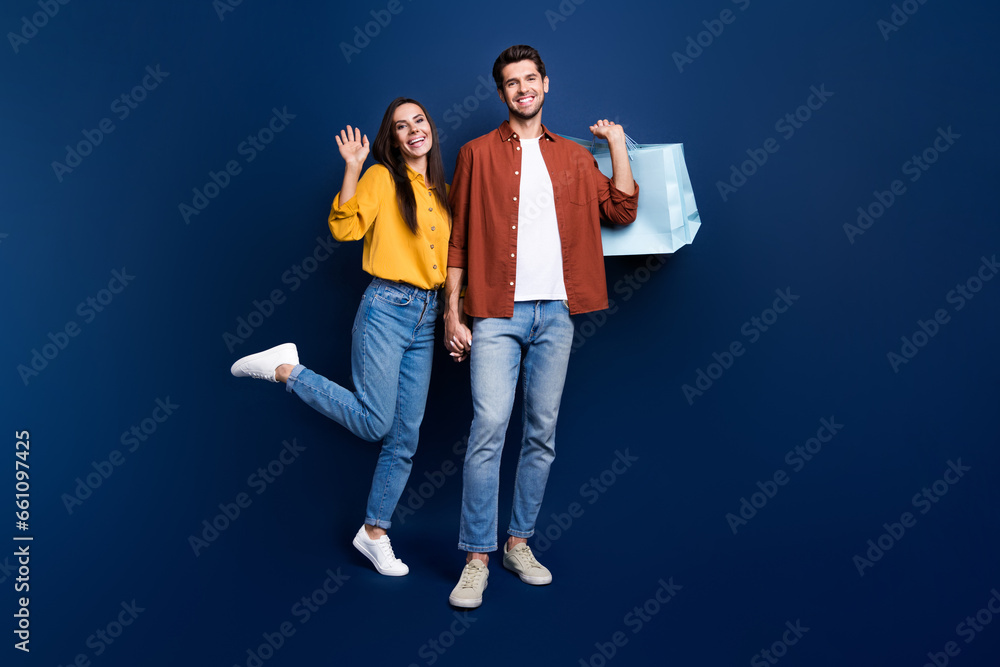 Full size photo of positive idyllic partners hold arms waving hi carry shop bags isolated on blue color background