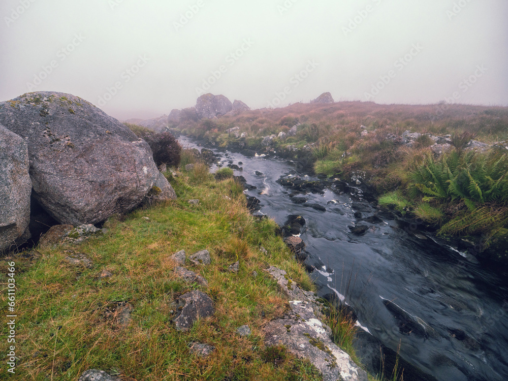 Obraz premium Irish nature scene with small creek in green fields with rocks and fog in the background. Connemara, Ireland. Hard weather conditions. Nature, travel and sightseeing.