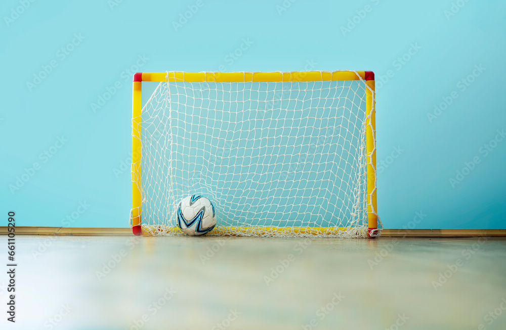 Fototapeta premium Football ball into soccer net of plastic children goal standing on wooden floor on playground inside