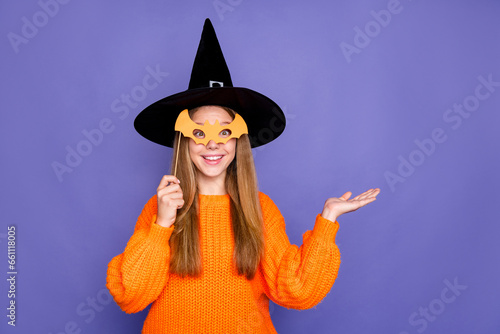 Photo of smiling devil witch teen girl wearing black cone hat holding mask showing brochure halloween isolated on violet color background