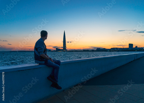 Alone man sitting with his back on embankment of river, canal and watching Skyscraper Lakhta center at sunset. White night. Concept think, reflection, achievement. Travel and tourism. Selective focus. photo