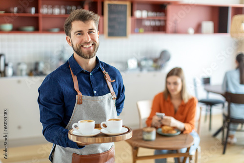 Cheerful male waiter carrying coffee cups on tray and smiling at camera, working in his small business restaurant cafe
